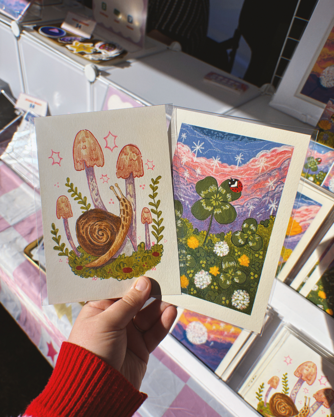 a hand holding 2 whimsical mixed-media art prints from the Nature of Belonging Collection by Luna Bramlett