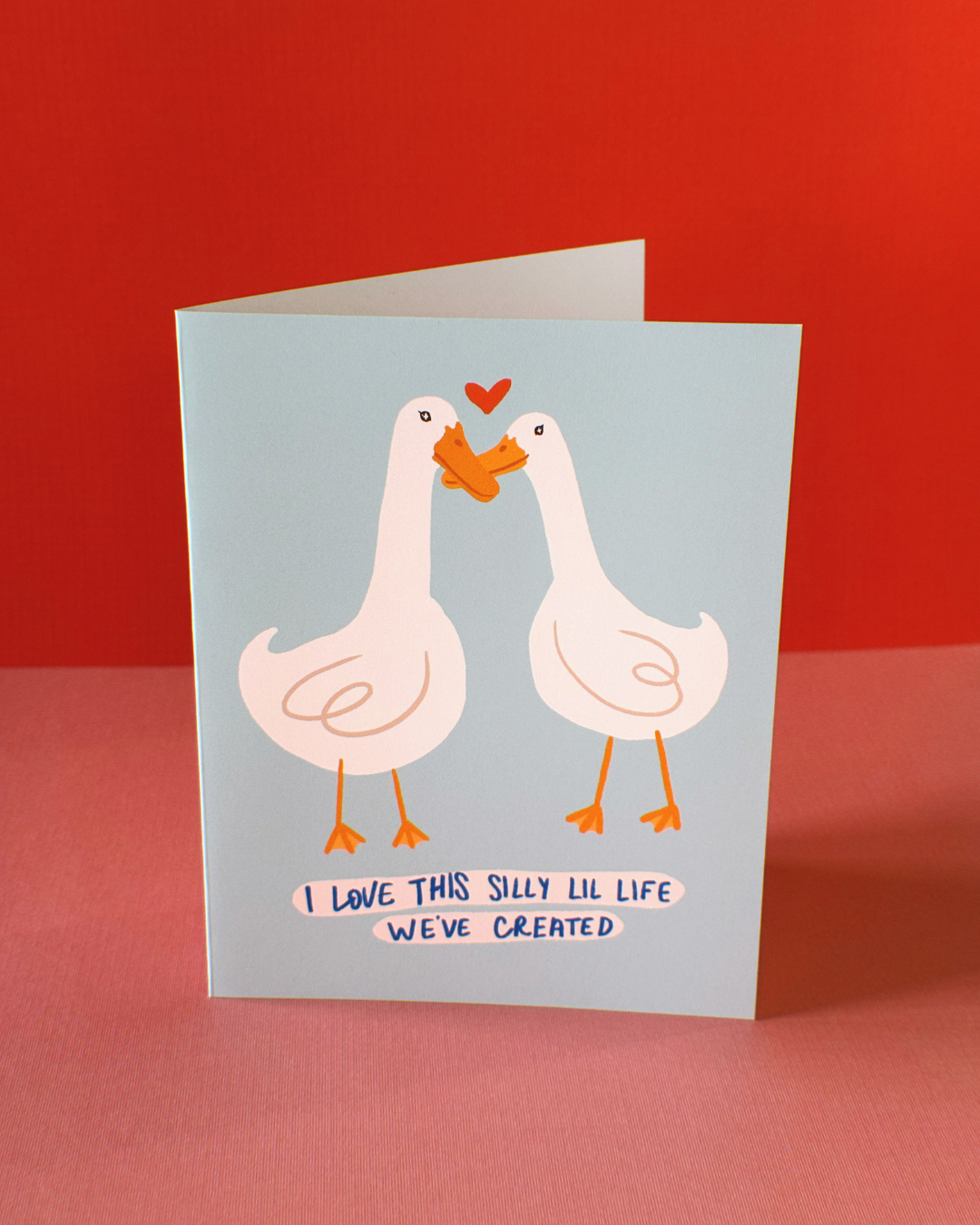 light blue greeting card with two white geese nuzzling each other's beaks with a heart above them. The hand written text says "I love this lil life we've created"