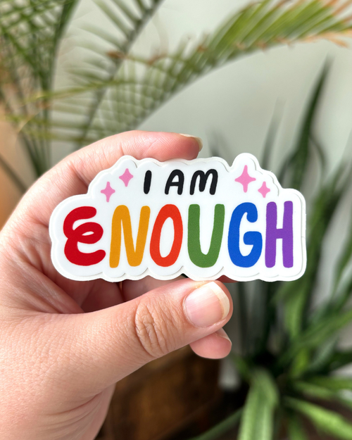Easy Peel 3in x 1.5in vinyl sticker with whimsical hand-lettered affirmation that says "I am Enough" on a white background. The letters "I am" are black with pink sparkles on either side, and they are stacked above the word "Enough." Each letter of the word is a color of the rainbow.