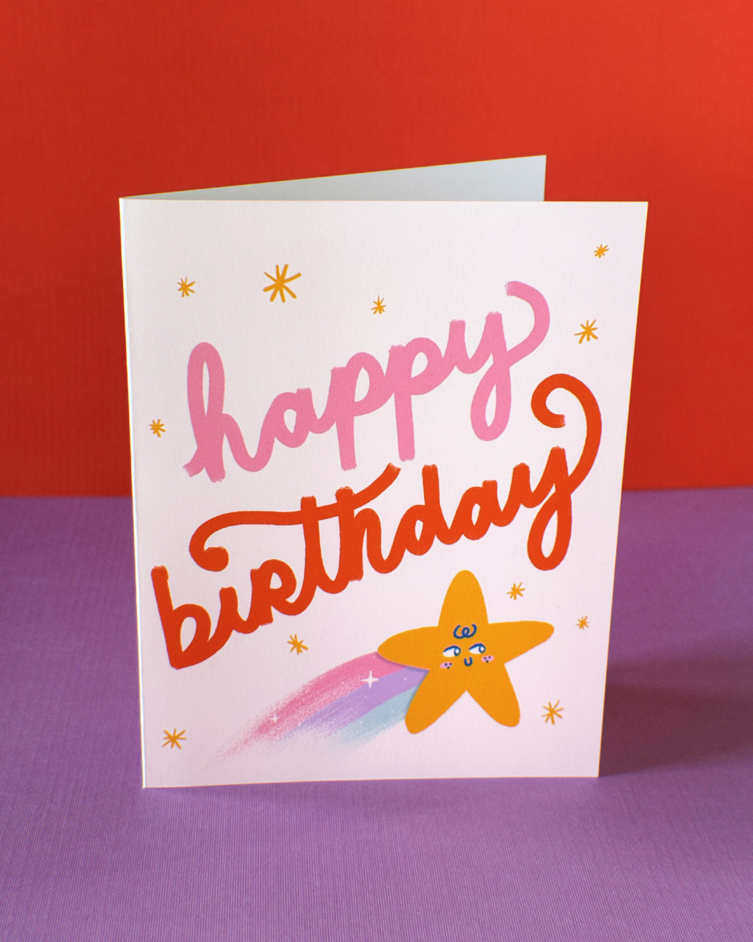 Cream colored greeting card that says happy birthday surrounded by small gold stars and one large gold shooting star with a cute face on it.