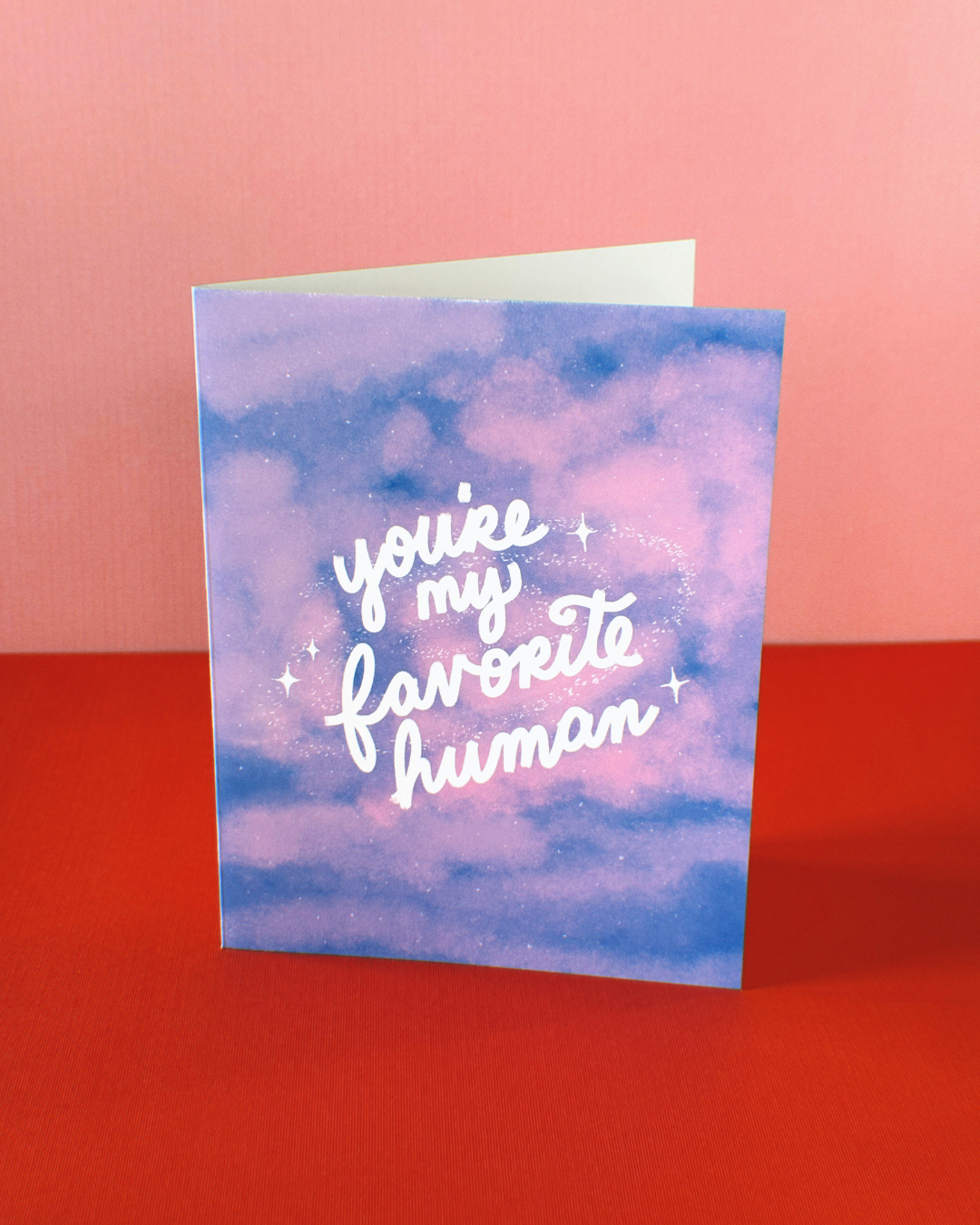 greeting card covered with pink clouds that says "You're my favorite human" in cursive surrounded by a subtle galaxy of stars